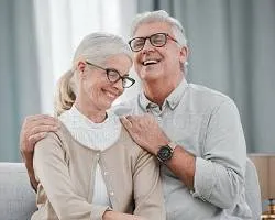 'A happy senior couple with grey hair and glasses smiling together, with the man gently holding the woman's shoulders.'