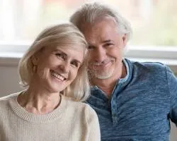 'Close-up portrait of a smiling middle-aged or senior couple leaning close to each other, with the woman on the left in a beige sweater and the man on the right in a blue shirt.'