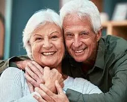 'Close-up of an elderly couple with white hair smiling together, with the man embracing the woman from behind and holding her hand.'